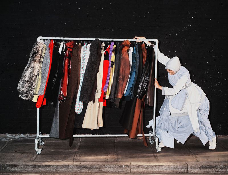 Adult female in layered clothing pushes a rolling rack of assorted coats indoors, against a dark grey wall and grey stone floor.