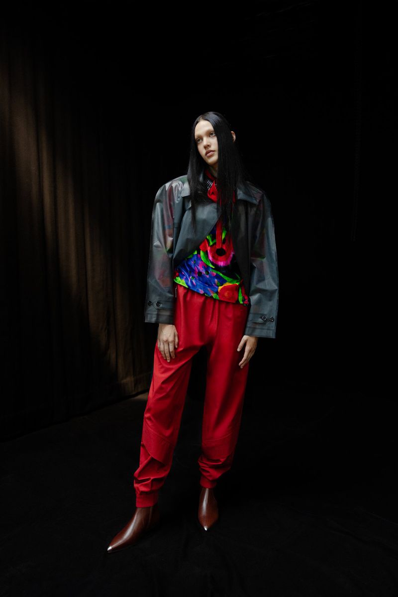 A model with long hair poses in a colorful floral top, gray jacket, and red pants against a dark background in a fashion-forward studio shot.