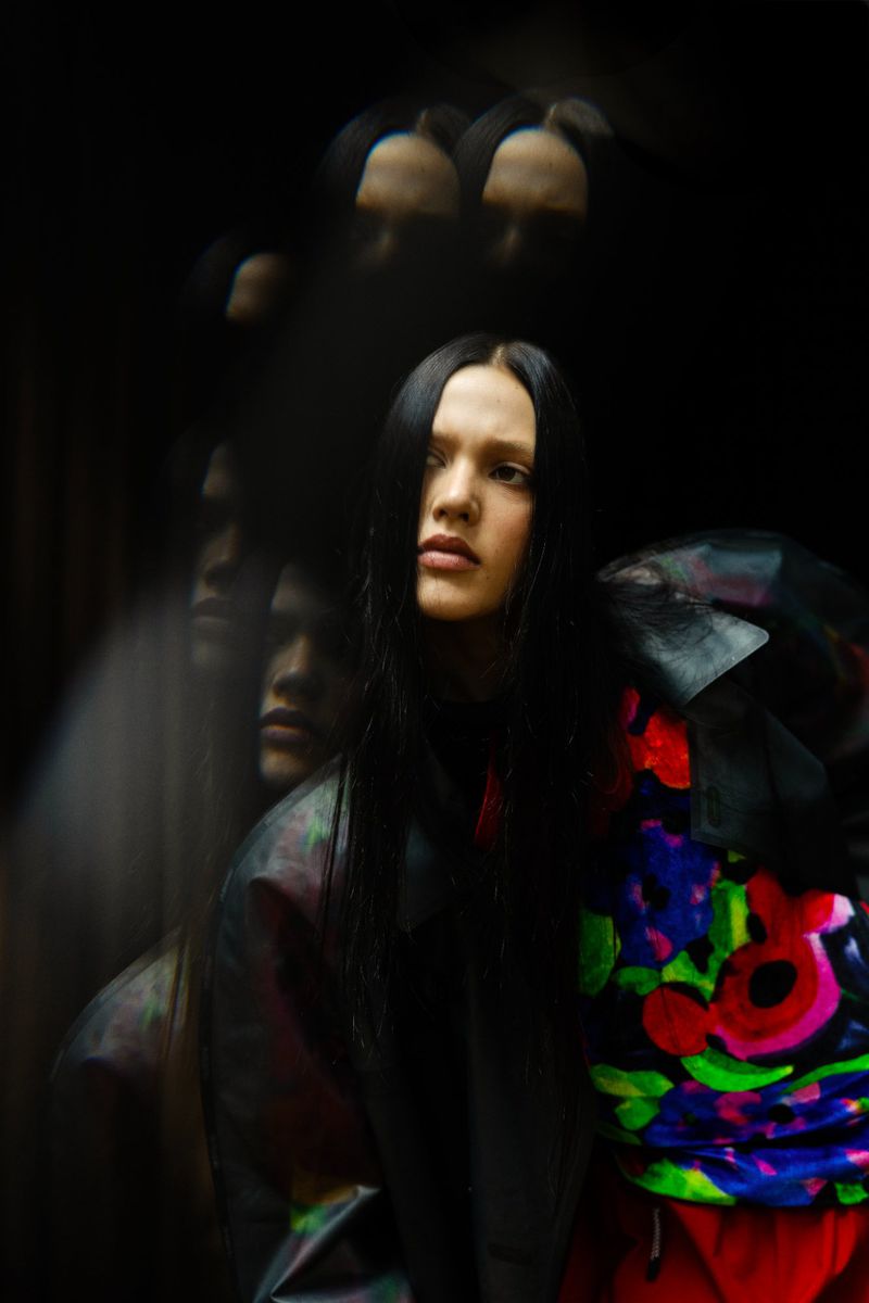 Portrait of a woman with long black hair, a black jacket, and a multi-colored shirt against a blurred dark background.