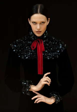 Elegant fashion portrait of a woman with fair skin and black hair wearing a black dress with a decorative collar and cuffs and a red bow.