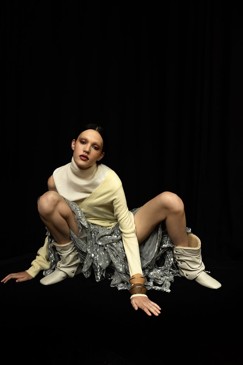 Fashionable adult female poses in a light yellow and silver dress with white boots in a studio setting against a black backdrop.