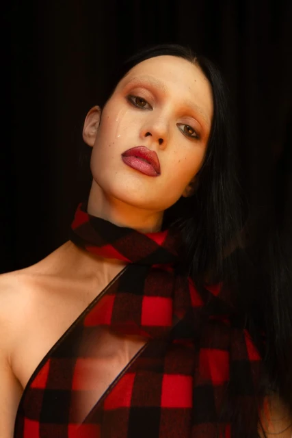 Close-up portrait of a woman with pale skin, dark hair, and a red and black checkered scarf. She has a tear running down her cheek.