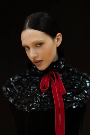 A striking portrait of a woman with dark hair, pale eyebrows, and a distinctive black ruffled collar with a bright red ribbon.