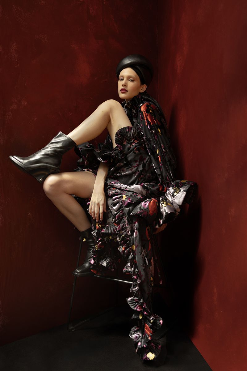 A woman in a floral dress and black boots poses in a studio with red walls, creating a dramatic and fashionable image.