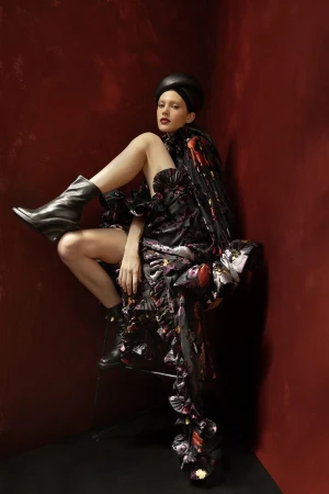 A woman in a floral dress and black boots poses in a studio with red walls, creating a dramatic and fashionable image.