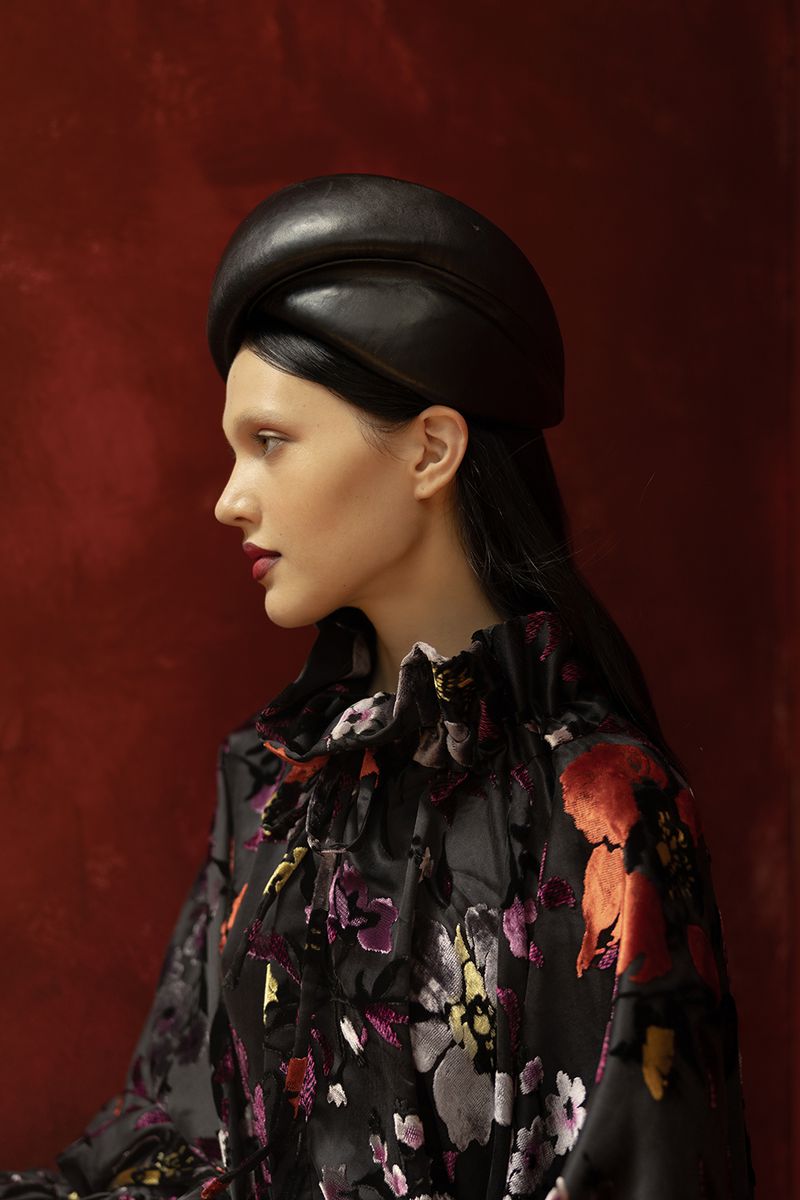 A model showcases a unique black leather hat and floral patterned top against a red textured backdrop in a studio fashion portrait.