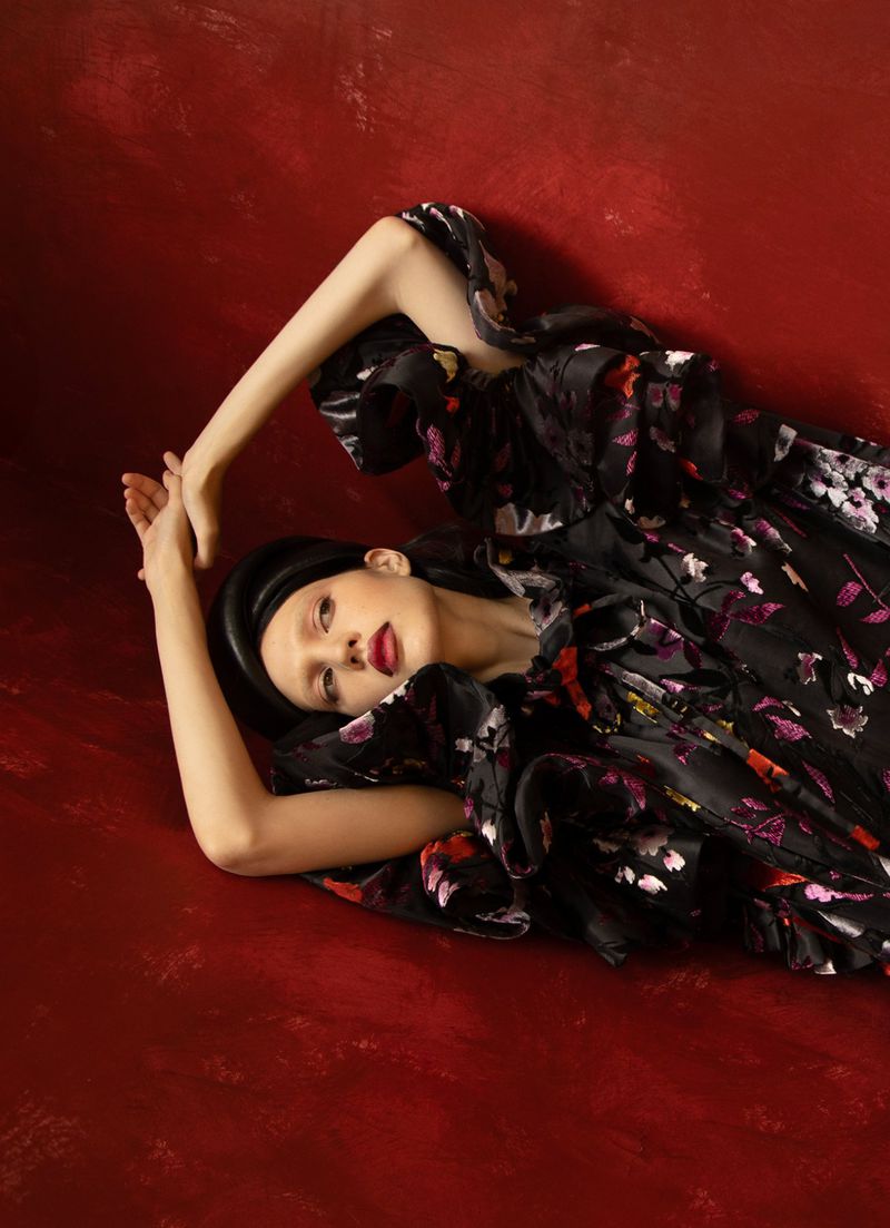 A woman wearing a black floral dress and a black hat is lying down on a red surface in a fashion editorial-style studio portrait.