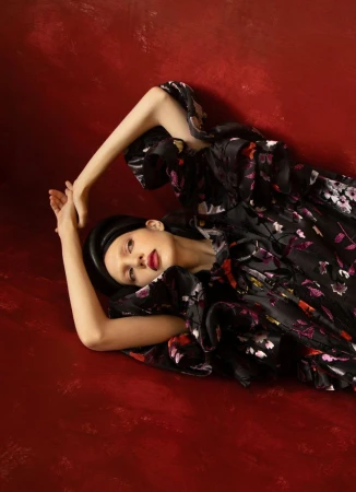 Model Abbey Fouts lying on a red textured surface wearing a voluminous black floral dress and padded black headpiece.