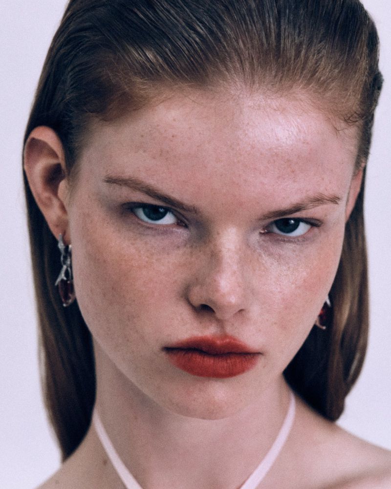 Close-up portrait of a fair-skinned woman with freckles and copper hair, wearing a simple top and minimal jewelry, against a plain background.