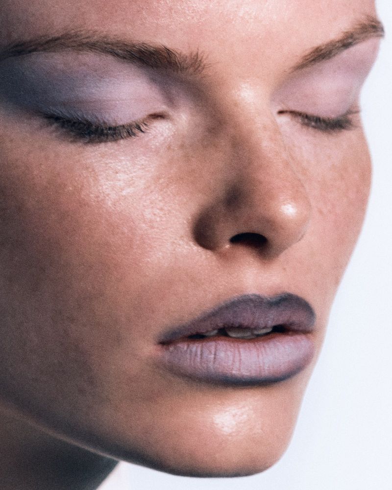 Close-up portrait of a woman with eyes closed, wearing lilac eye shadow and lipstick, set against a white background in a beauty shot.