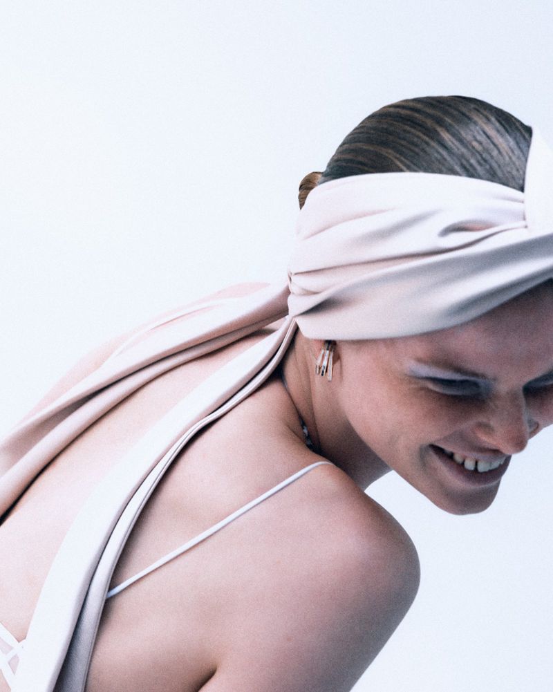 Smiling woman with bare shoulders, a light-pink satin wrap, and delicate earrings against a bright-white backdrop in a medium shot.