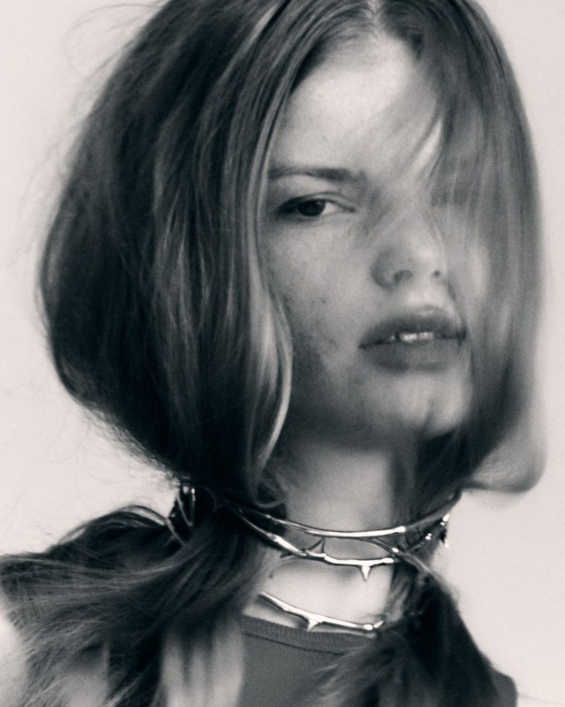 Close-up portrait of a young woman with dark blonde hair wearing a silver choker with spike-like details in a blurred monochromatic shot.