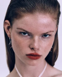 Close-up beauty portrait of model Elise Swain with freckles, slicked-back hair, and red lipstick looking directly at the camera.