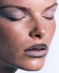 Close-up beauty portrait of a model with pale purple eyeshadow and dark lip liner against a plain white background.