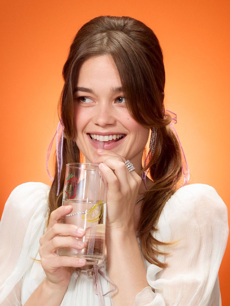 Smiling woman with pigtails drinks water through a straw from decorated glass. Bright orange background. Close-up studio shot.