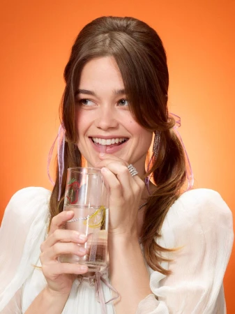 Smiling woman with pigtails drinks water through a straw from decorated glass. Bright orange background. Close-up studio shot.