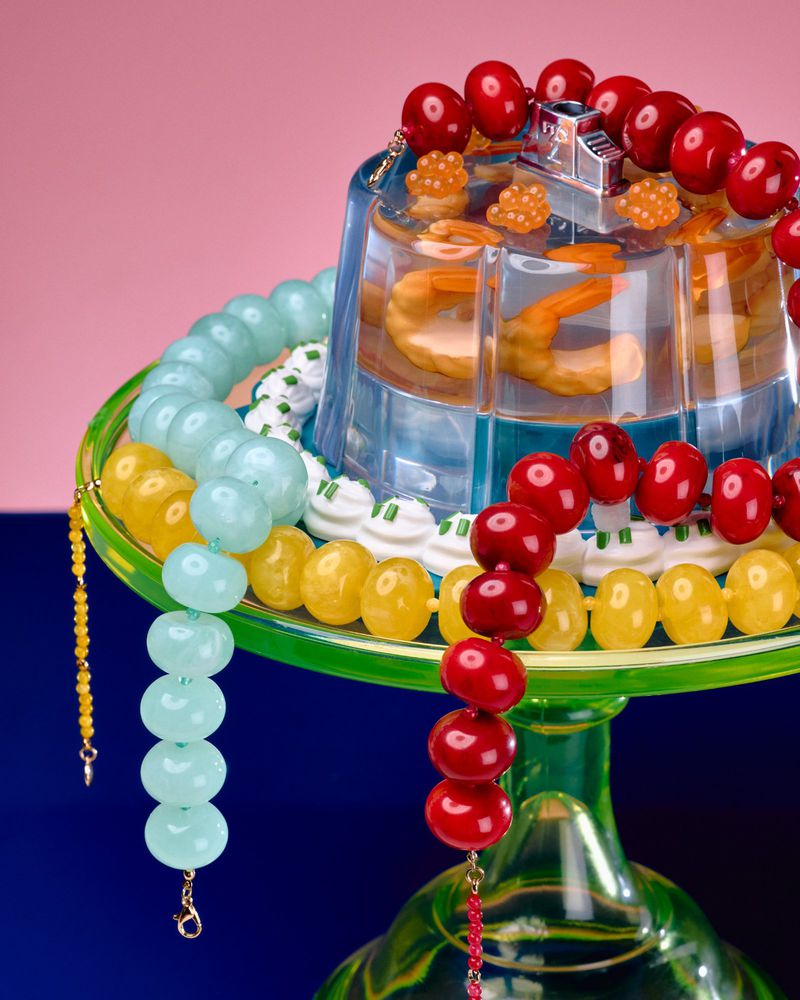 Elevated arrangement of food-filled plastic molds encased in bead necklaces on a green glass stand against a split pink and blue backdrop.