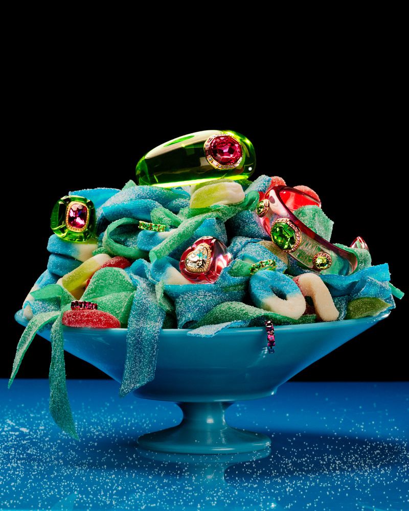 An overflowing bowl of gummy candies and costume jewelry on a blue surface against a black backdrop, creating a vibrant and whimsical scene.
