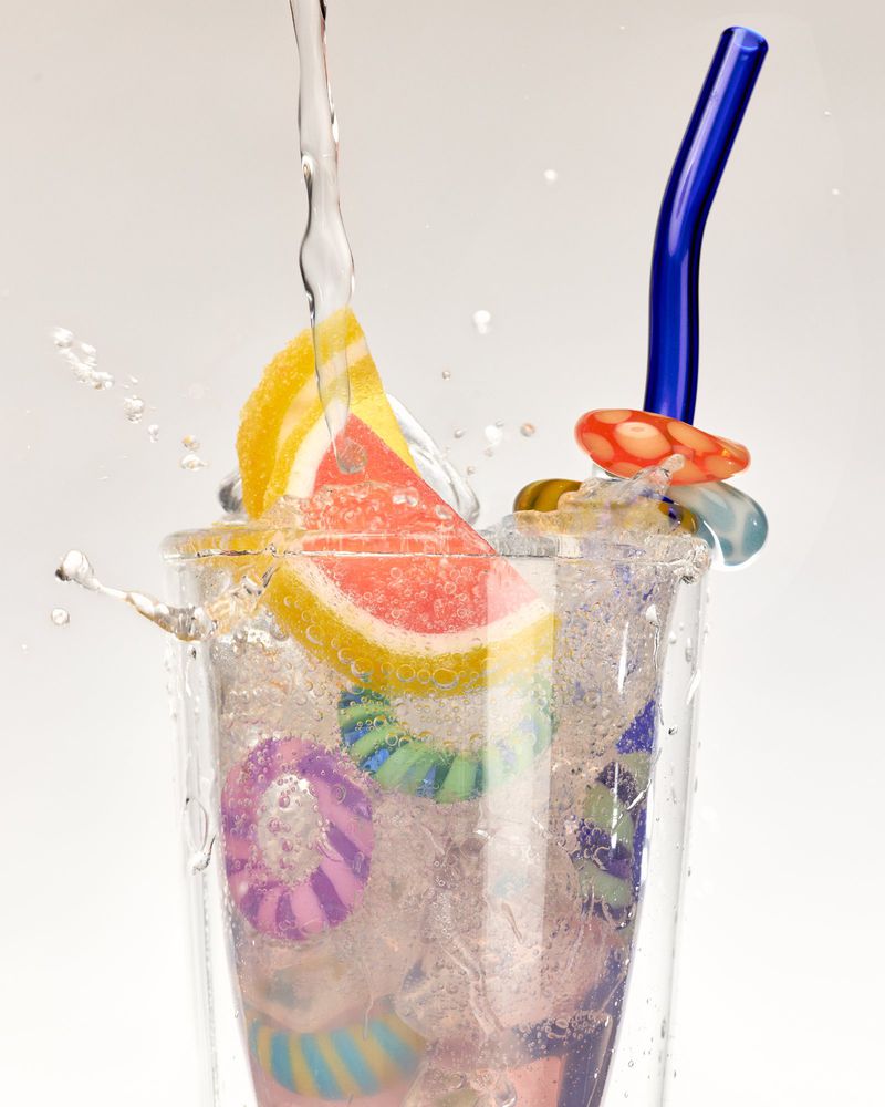 Refreshing glass of water being poured with fruit and colorful candy rings, blue drinking straw, and a plain white backdrop.