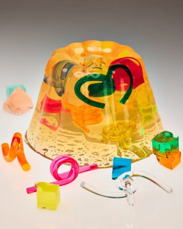 Molded translucent yellow gelatin cake containing suspended colorful twisted acrylic shapes surrounded by geometric cubes on a light surface.