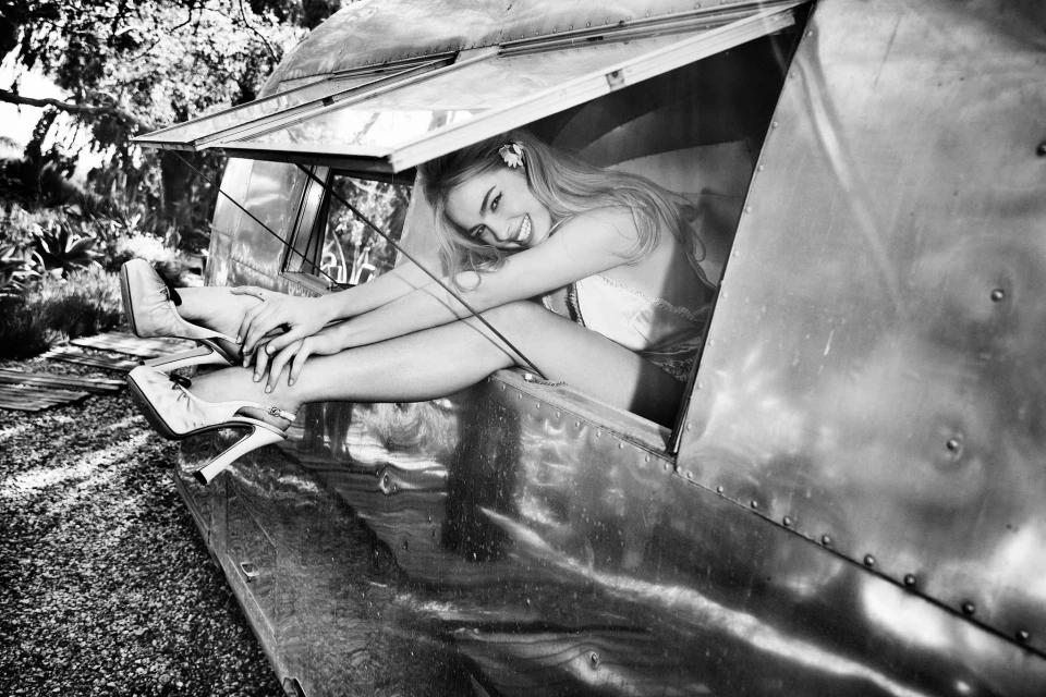 Black and white shot of a smiling young woman sitting inside an aluminum trailer with legs extended wearing high heels.
