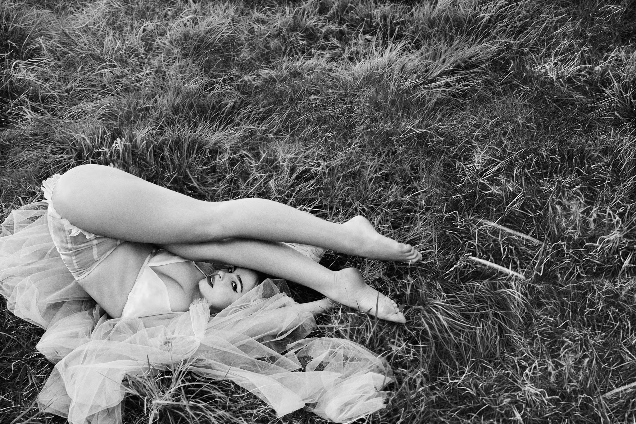 Black and white photograph of a woman lying in the grass, wearing lingerie and sheer fabric, in an outdoor setting.