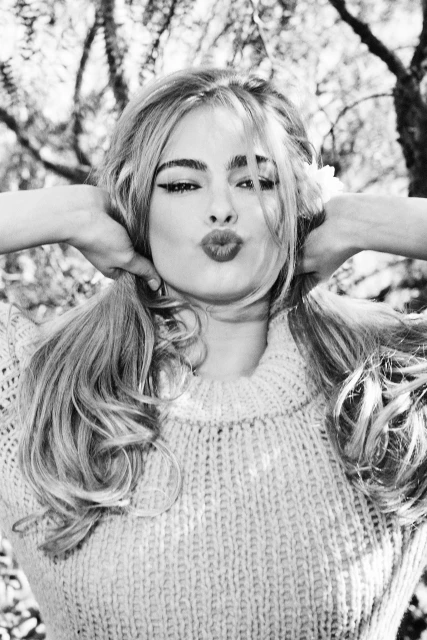 Black and white portrait of a woman with long curly hair and a sweater, with her hands behind her head and making a kissy face.