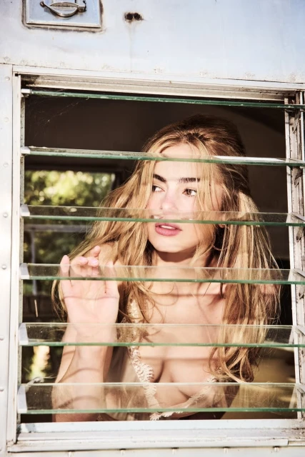 A blonde woman peers through the blinds of a vintage silver trailer, soft light illuminating her face in this medium shot.
