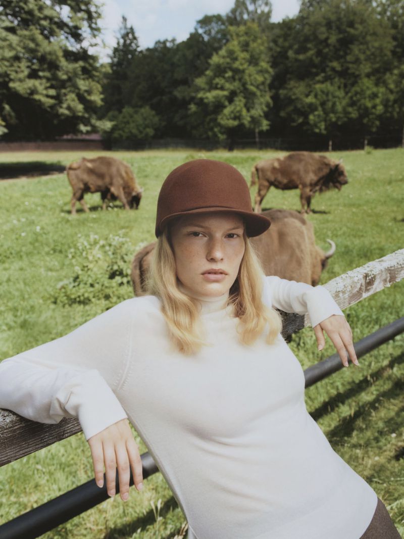 A young adult woman in a white sweater and brown cap leans against a fence in a green field with European bison in the background.