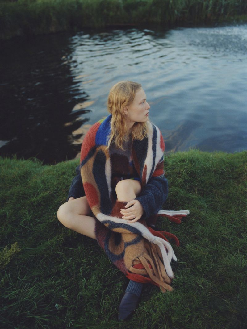 Young woman in a patterned shawl sitting cross-legged on a grassy riverbank looking off to the side in natural light.