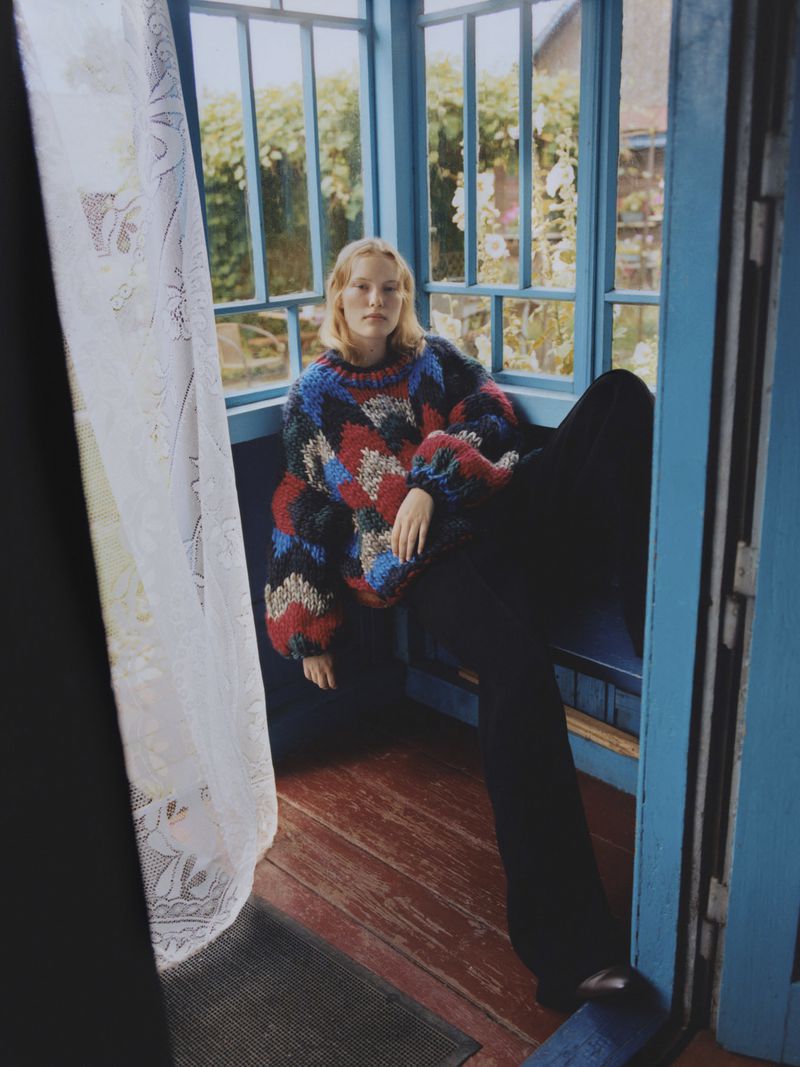 A young woman with blonde hair is seated in a sunroom, wearing a colorful patterned sweater and dark pants, illuminated by natural light.