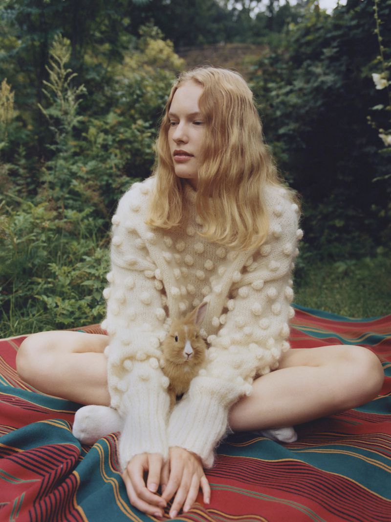 Young woman in white sweater with small rabbit on lap sits cross-legged on striped blanket in sunny outdoor grassy setting.