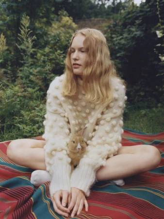 Young woman in white sweater with small rabbit on lap sits cross-legged on striped blanket in sunny outdoor grassy setting.