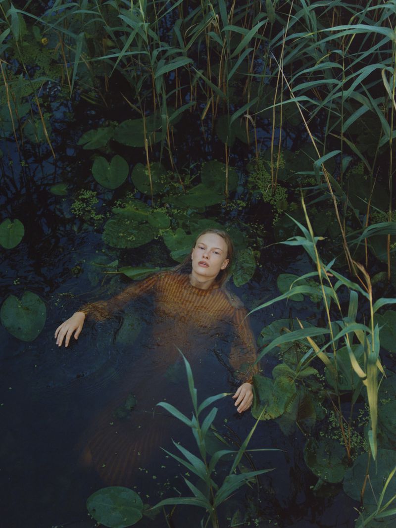 Woman in brown sweater submerged in dark water surrounded by lily pads and tall green reeds in a natural outdoor environment.