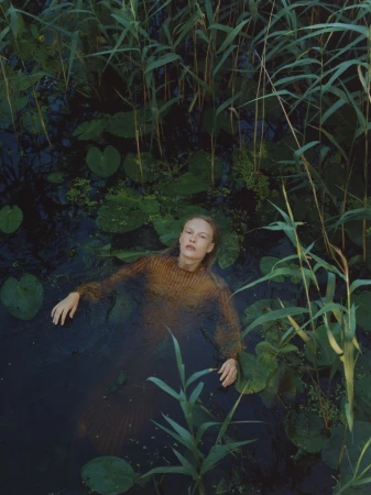 Woman in brown sweater submerged in dark water surrounded by lily pads and tall green reeds in a natural outdoor environment.