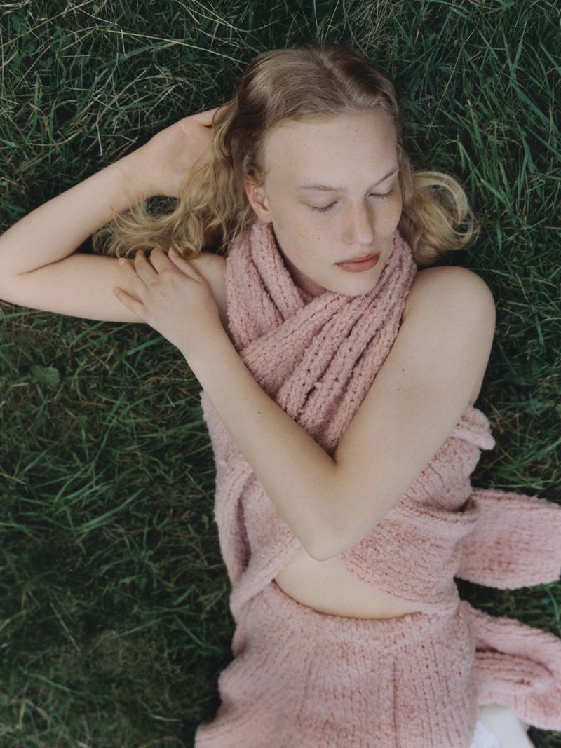 Overhead shot of a fair-skinned adult woman with wavy blond hair lying in green grass wearing a pink knit outfit.
