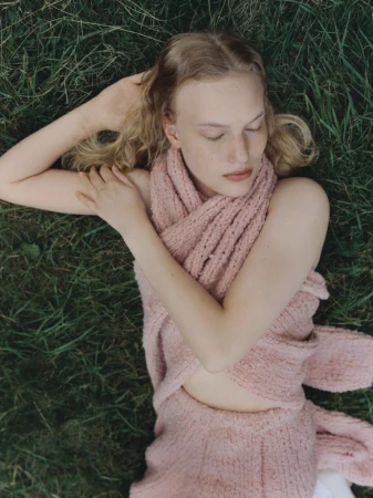 Overhead shot of a fair-skinned adult woman with wavy blond hair lying in green grass wearing a pink knit outfit.