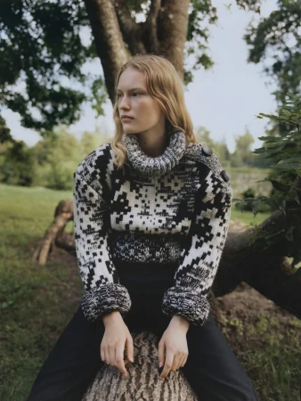 Model Astrid Østergaard sitting on a fallen tree trunk wearing a patterned black and white turtleneck knit sweater outdoors.