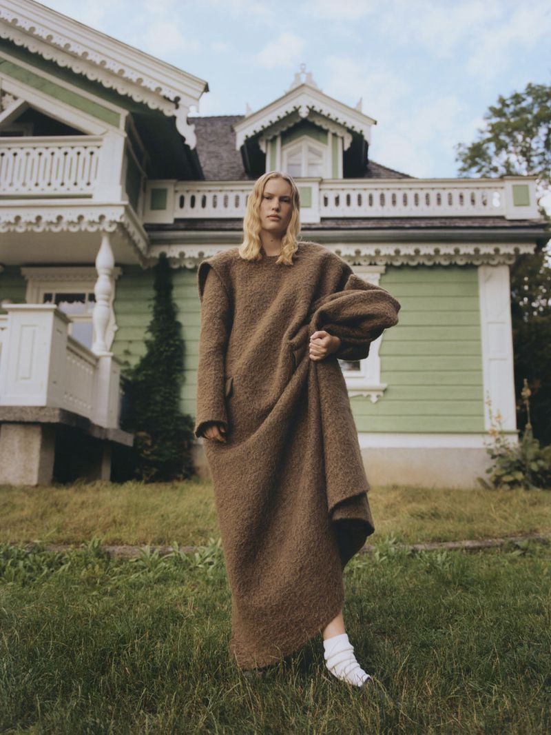 An adult female fashion model poses in a long brown garment on a lawn in front of a green and white wood house on a cloudy day.