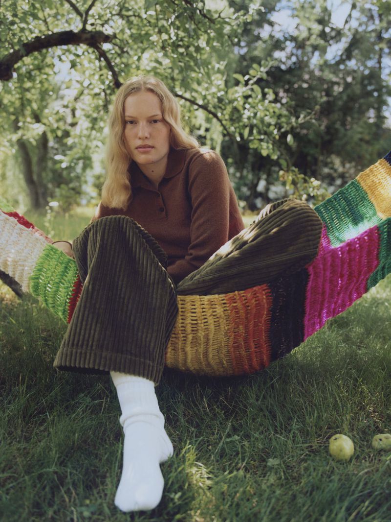 Woman in brown top and corduroy pants relaxes in a colorful hammock on green grass under a tree in natural light.