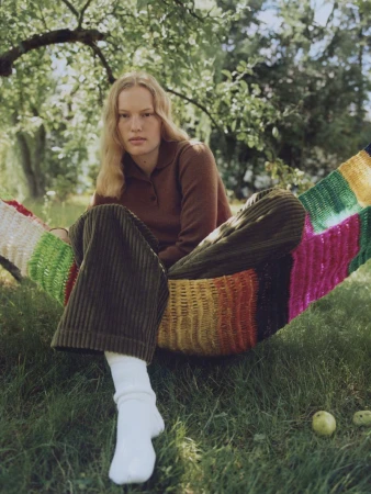 Woman in brown top and corduroy pants relaxes in a colorful hammock on green grass under a tree in natural light.
