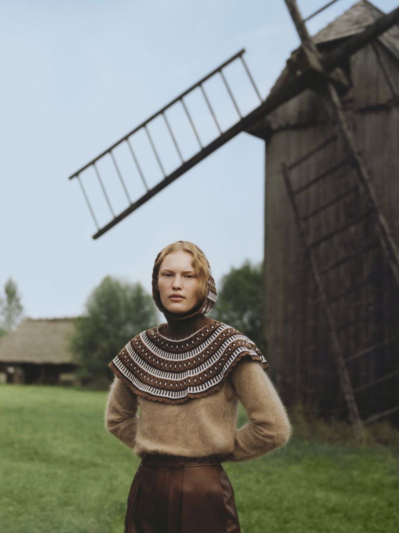 A woman in a brown sweater and bonnet poses in front of a large windmill and a small building in an outdoor grassy setting.