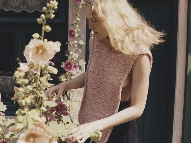 Woman in sleeveless top stands amongst pink hollyhock flowers against the exterior wall of a building in natural daylight.