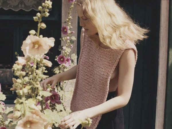 Woman in sleeveless top stands amongst pink hollyhock flowers against the exterior wall of a building in natural daylight.