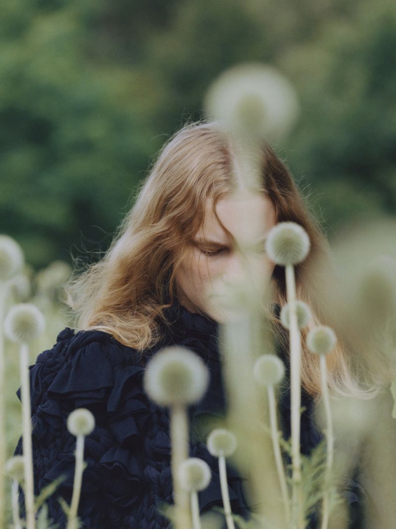 Portrait of a young woman with long blond hair wearing a black dress, standing outdoors in a field of globe thistle flowers.