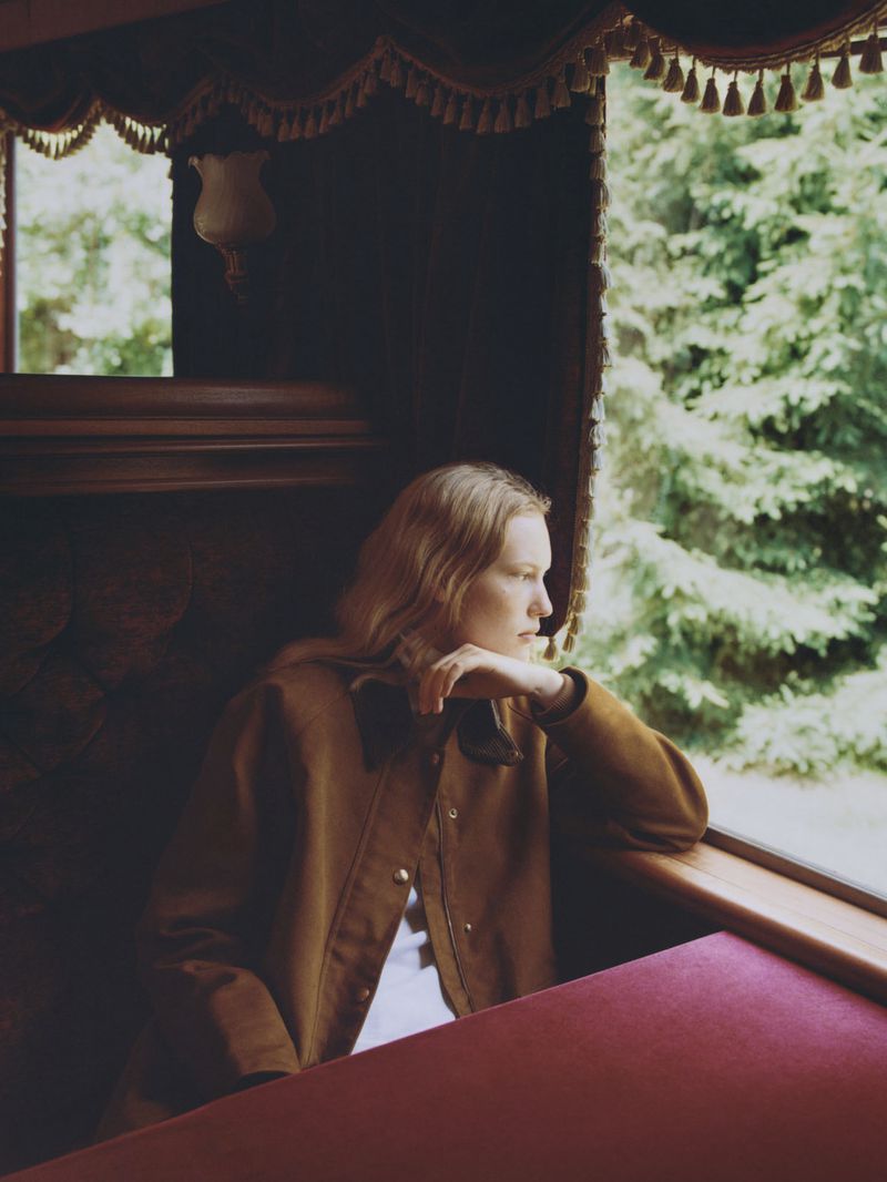 Woman in brown jacket sits in vintage train car, gazing out window at a forest. Moody lighting, muted colors, travel, and fashion themes.