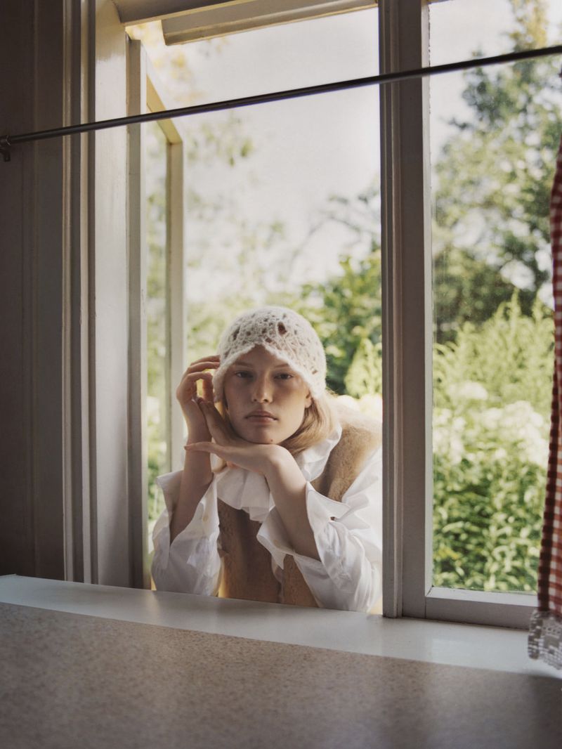 A young woman with light skin and blonde hair stands inside a white-framed window wearing a white lace hat and ruffled white shirt.