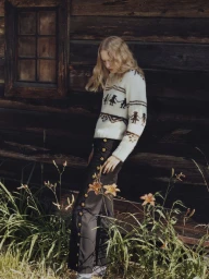 Model Astrid Østergaard wearing a patterned cream knit sweater and dark trousers leaning against a dark wood cabin exterior.