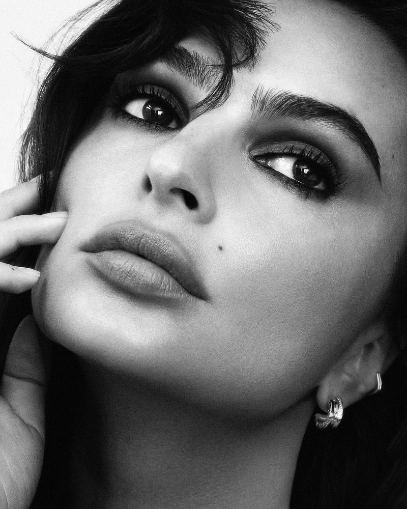 Monochrome close-up portrait of Emily Ratajkowski with dark eye makeup and hoop earrings against a neutral background.