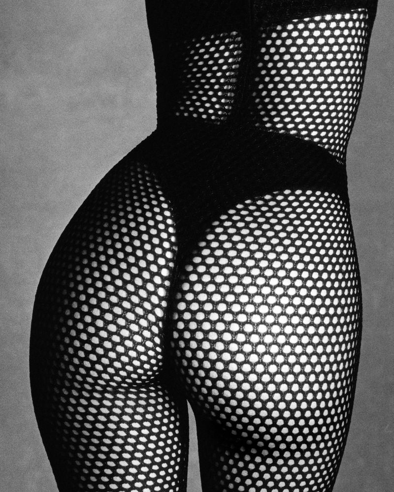 Close-up studio shot of a Black adult female torso and legs wearing a fishnet bodysuit against a grey backdrop with soft lighting.
